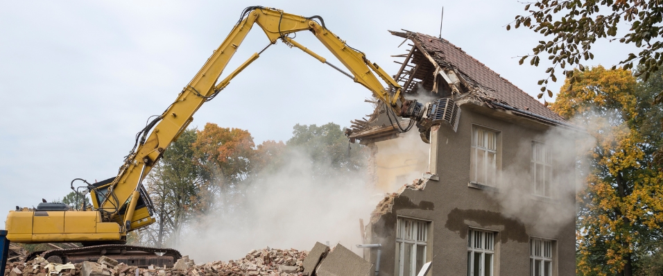 Excavator performing house demolition with debris and dust in the foreground.